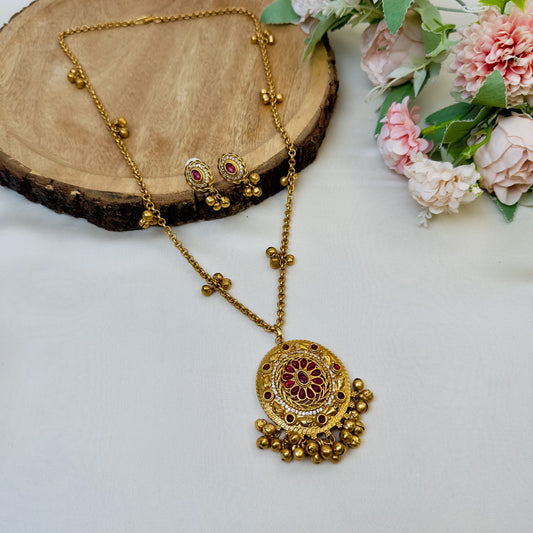 A gold-colored Ghungroo Mala Pendant Set with intricate designs and ornamental details, displayed on a wooden surface with flowers in the background.
